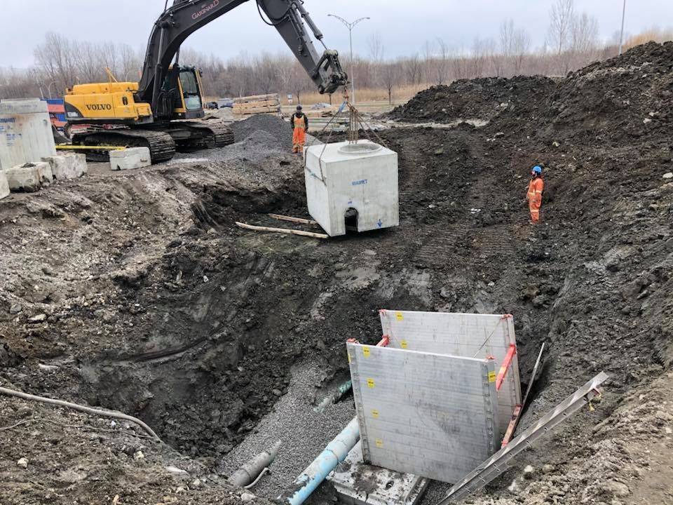 Une excavatrice soulève une structure en béton et la dépose dans une fosse de construction. Des ouvriers en gilets orange supervisent la manœuvre.