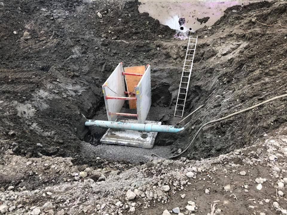 Fosse d'excavation avec tuyau, étaiement, échelle et base en béton dans un sol boueux.