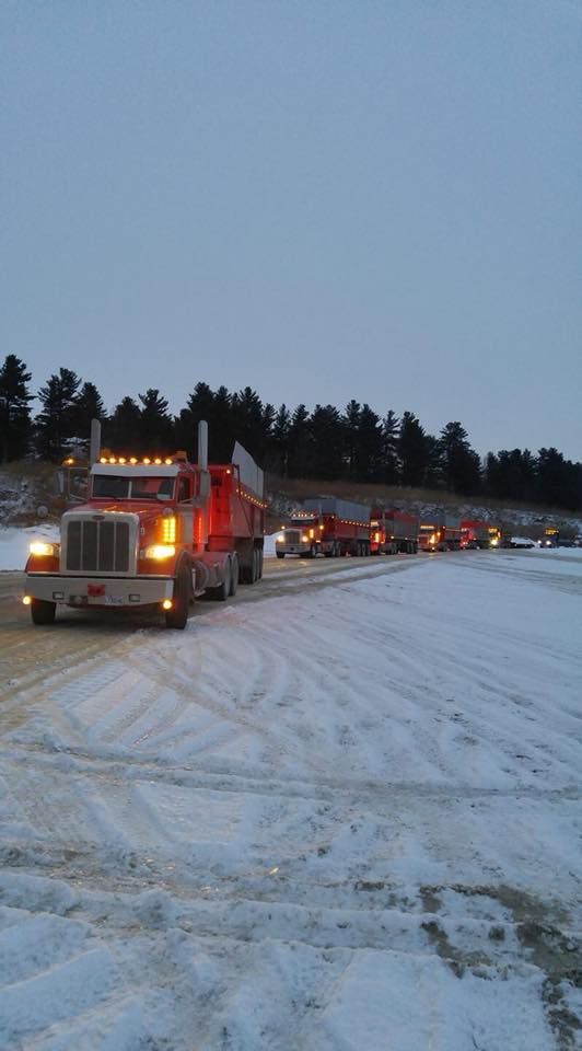 Une file de camions-bennes orange sur une route enneigée, éclairés par des phares, sous un ciel crépusculaire.