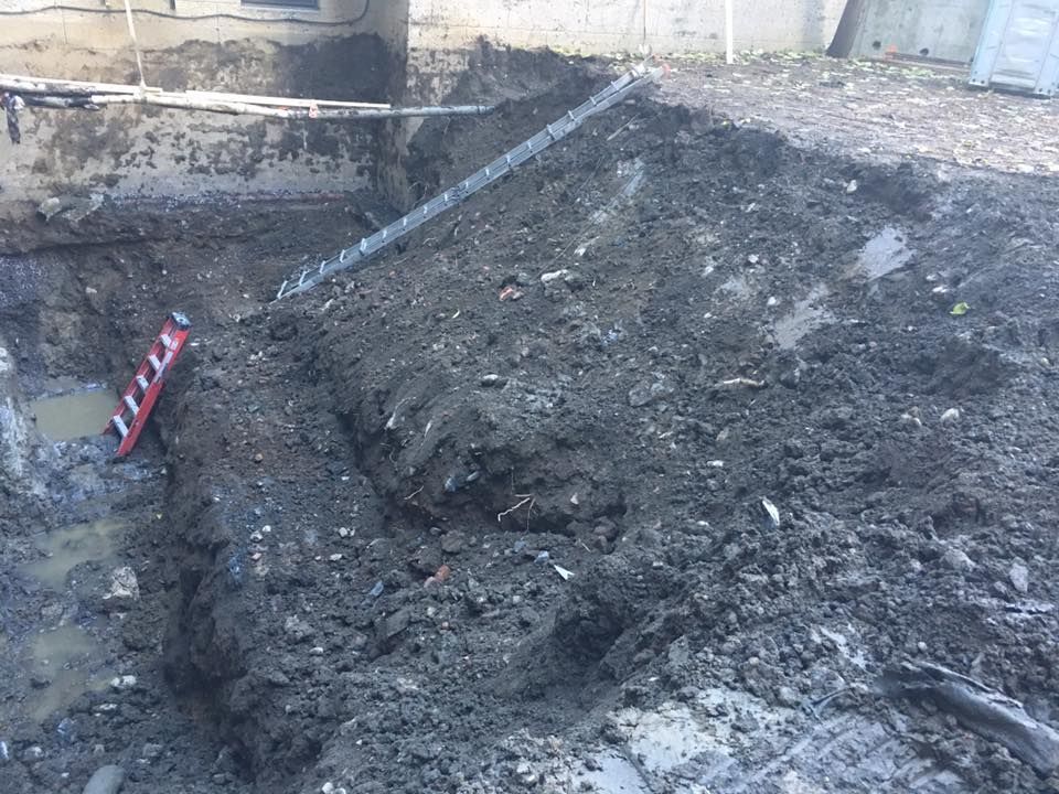 Un chantier de fouilles boueux jouxte un bâtiment. Un niveau gît dans la tranchée.