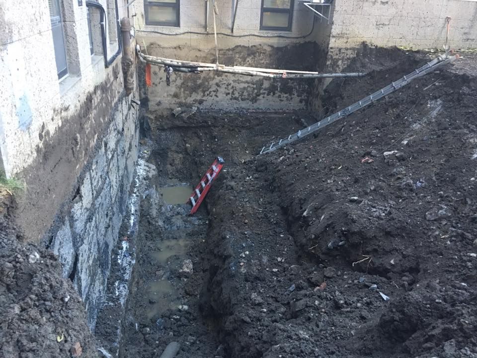 Travaux d'excavation à proximité d'un bâtiment. Terre, échelle et mur en béton visibles.