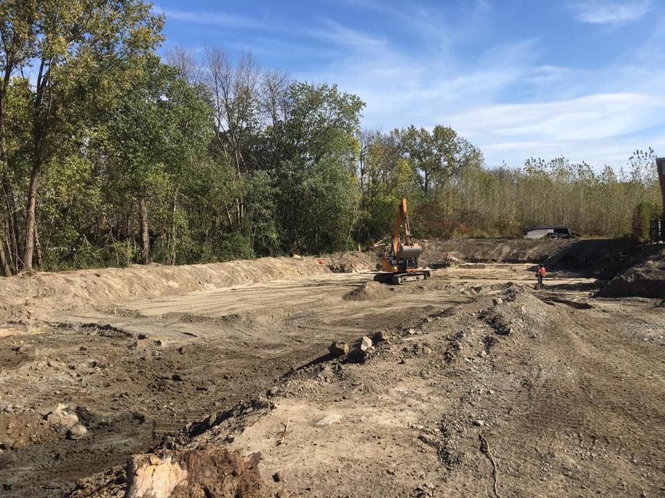 Chantier avec excavatrice. Terre et arbres sous un ciel bleu partiellement nuageux.