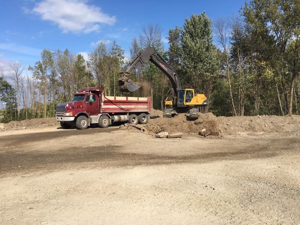 Une excavatrice charge de la terre dans un camion-benne rouge sur un chemin de terre, avec des arbres en arrière-plan sous un ciel bleu.