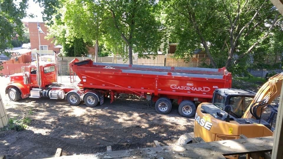 Camion-benne rouge et remorque sur un chantier, à côté d'une pelle mécanique Case. Journée ensoleillée.