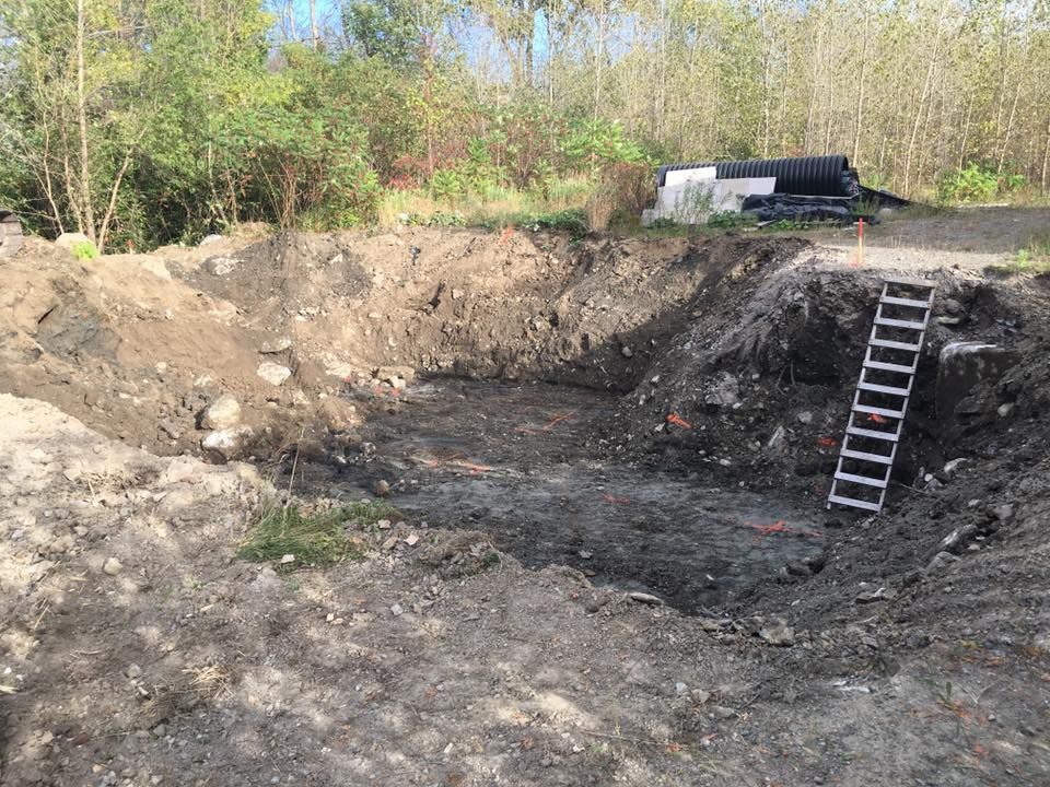 Grande fosse carrée creusée, aux parois de terre, avec une échelle et des arbres en arrière-plan.