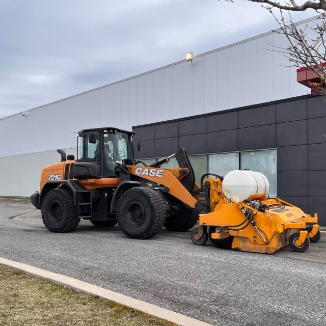 A bright orange Case front-end loader attached to a street sweeper attachment parked in front of a commercial building.