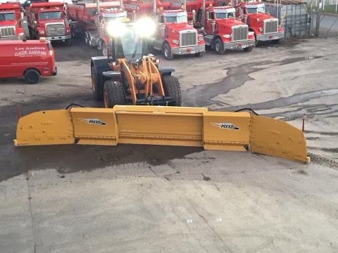 Front loader with yellow blade plowing asphalt, trucks in background.