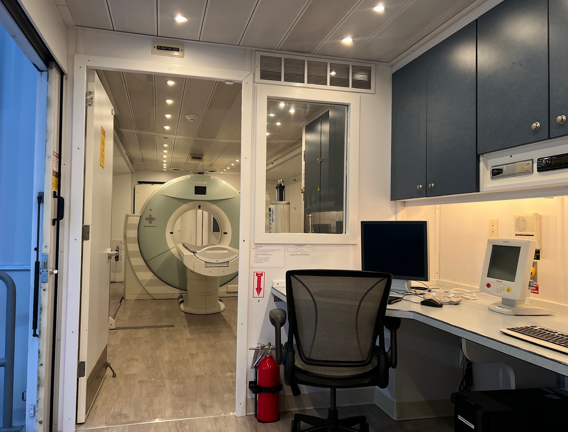 MRI control room with desk, monitor, chair, and view of the scanner through a window