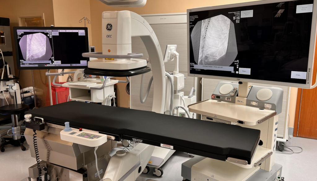 Hospital procedure room with a black operating table and two large monitors showing X-ray images.