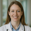 Smiling doctor in a white coat and blue shirt, standing in a clinic hallway.