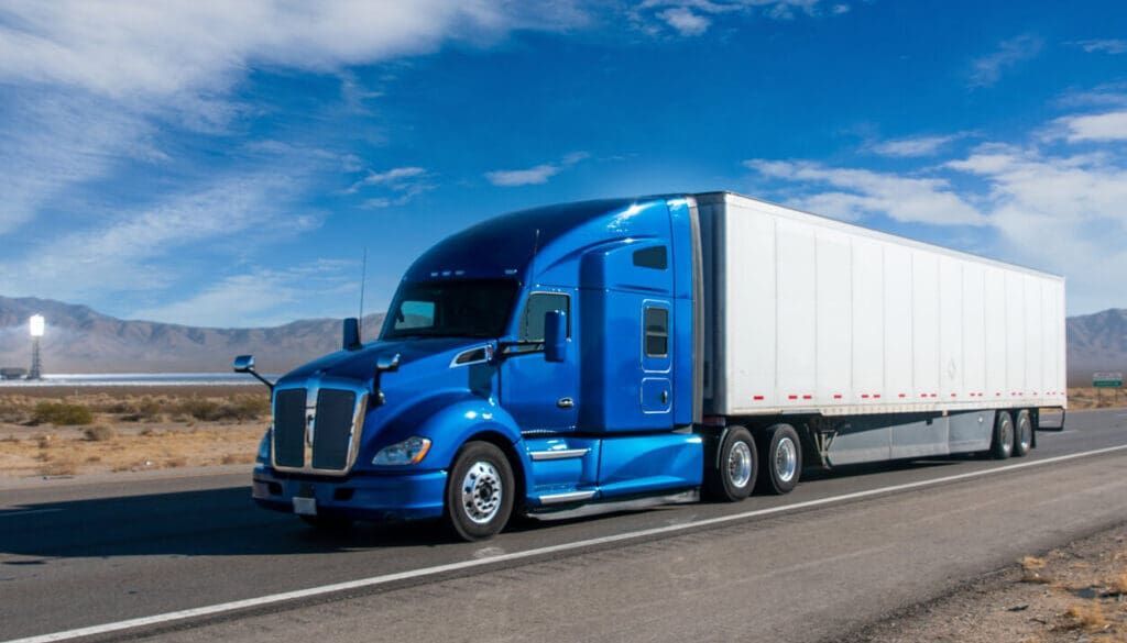 Blue semi-truck hauling a white trailer on a highway under a partly cloudy sky