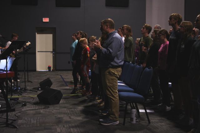 People singing and clapping in a dimly lit room, with a band on stage.