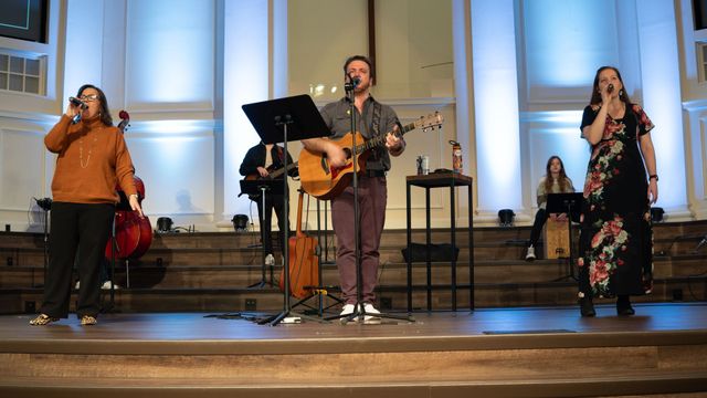 A musical group performs on a stage in a church. Two singers and a guitarist are in front; others in the background.