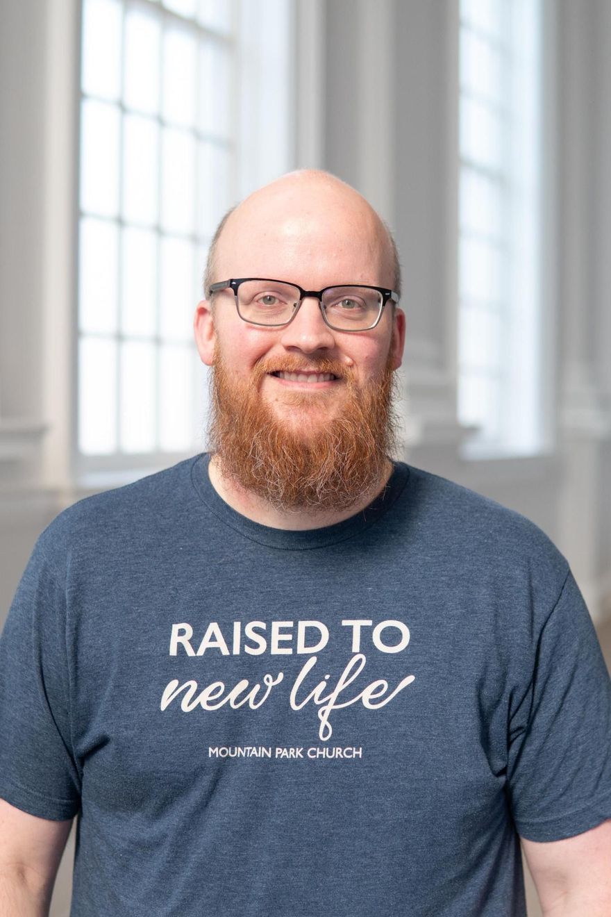 Man with a red beard and glasses smiles, wearing a gray shirt, neutral background.