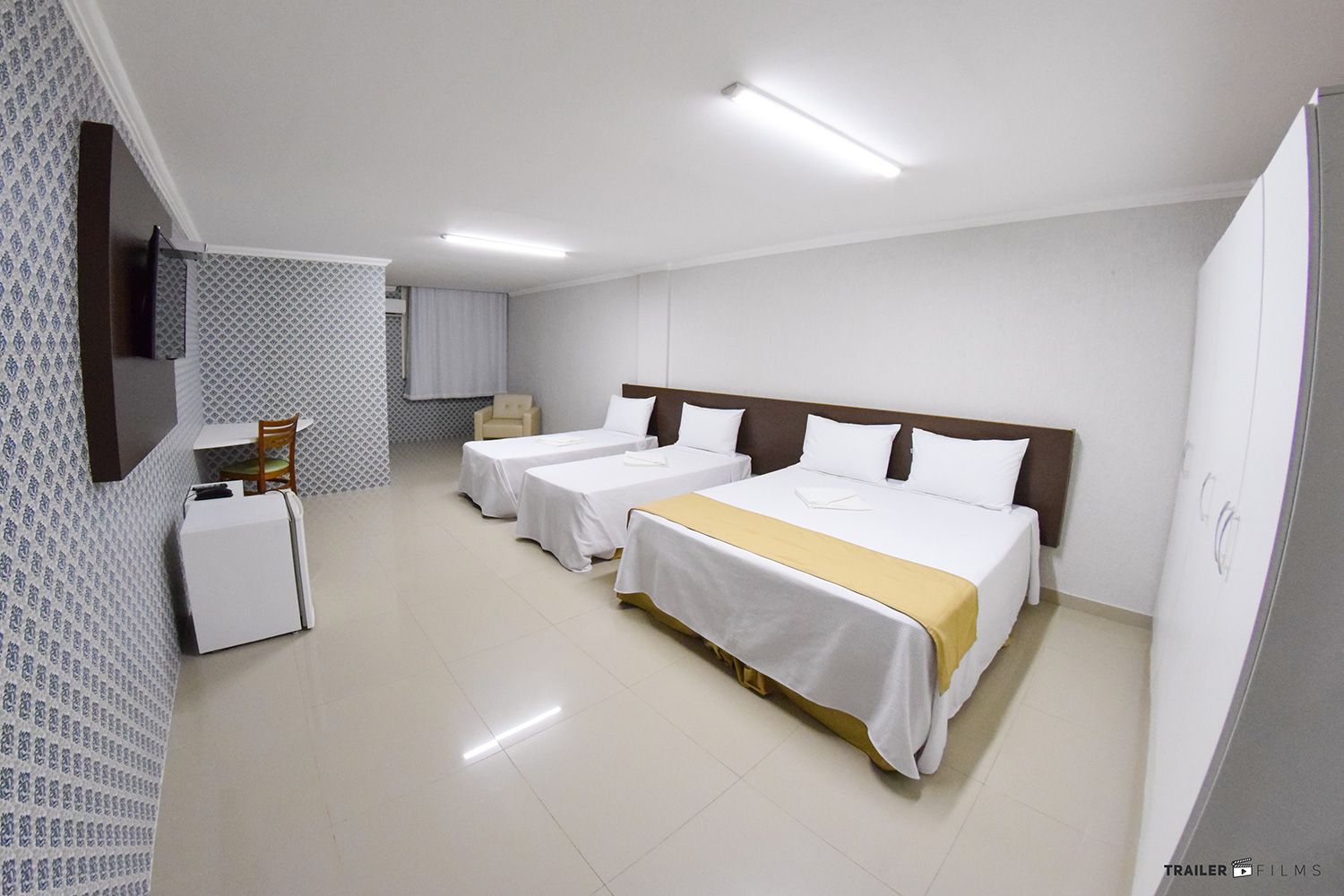 Quarto de hotel com três camas, uma escrivaninha, um guarda-roupa e uma parede com estampa decorativa.