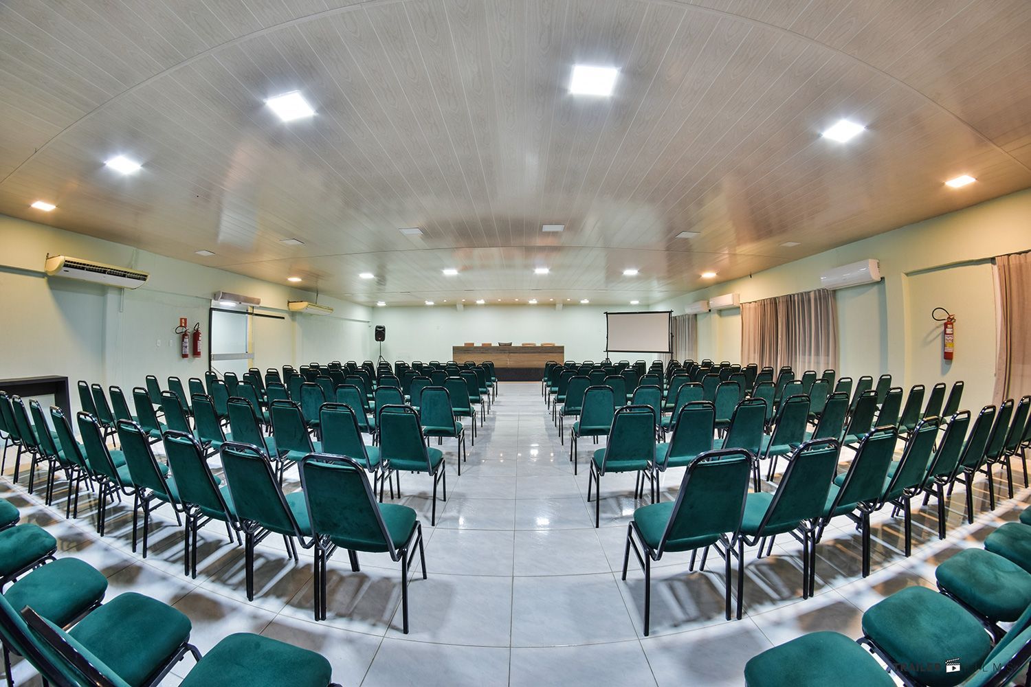 Sala de conferências vazia com cadeiras verde-azuladas dispostas em semicírculo de frente para um palco com tela de projeção.