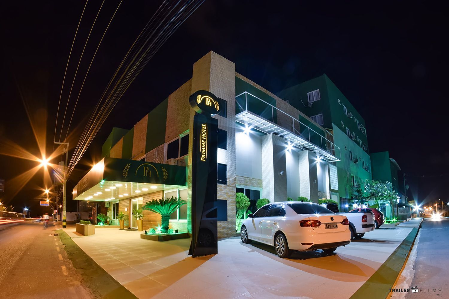Exterior do hotel à noite, fachada verde e branca com letreiro iluminado. Carros estacionados em frente.