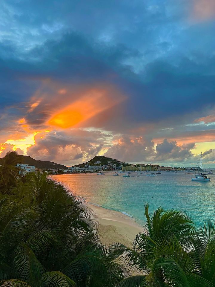 Sunset over a bay with boats. Orange and pink sky, blue water, green trees, and a sandy beach.