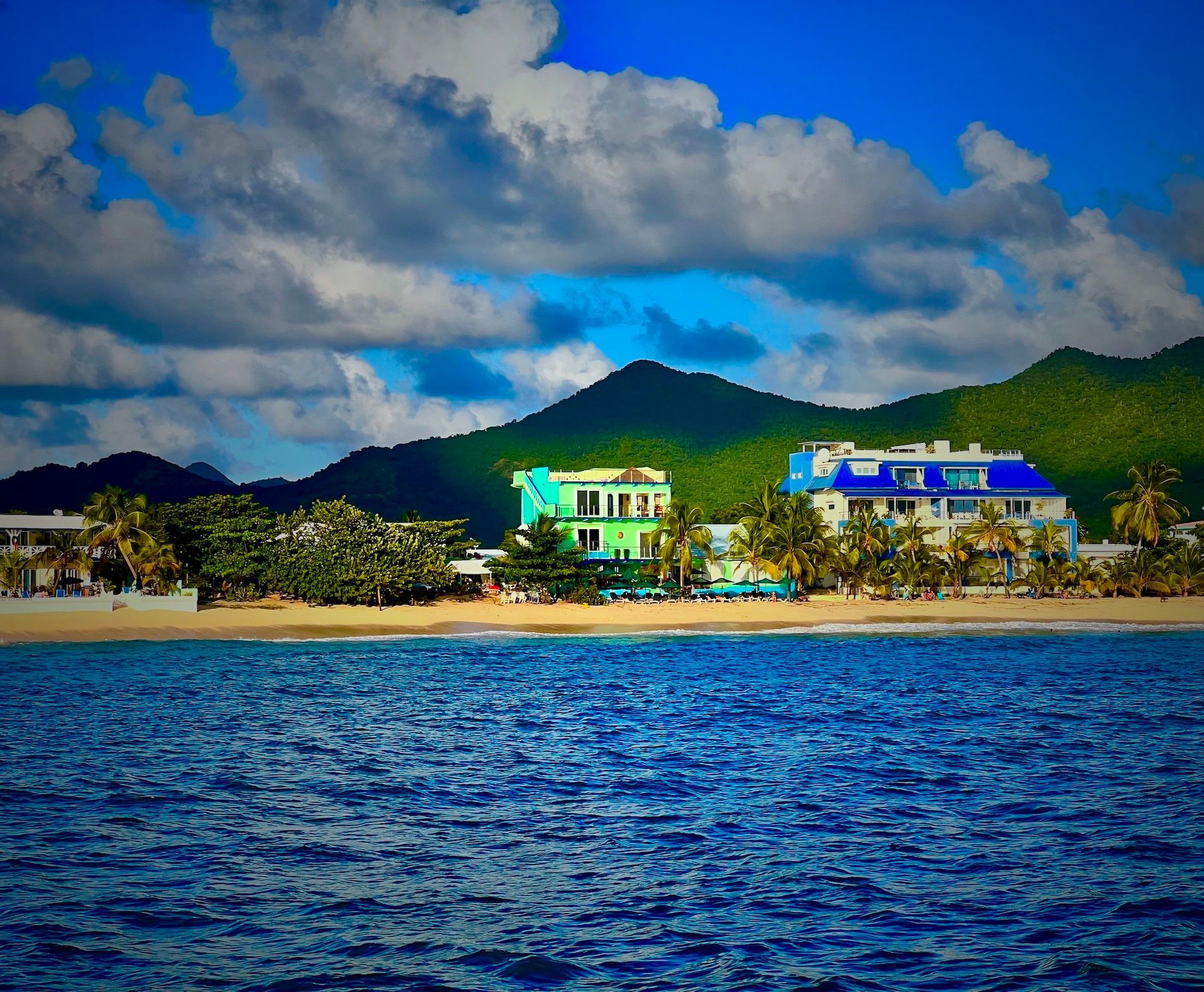 Ocean view with a sandy beach, buildings, green mountains, and a cloudy blue sky.