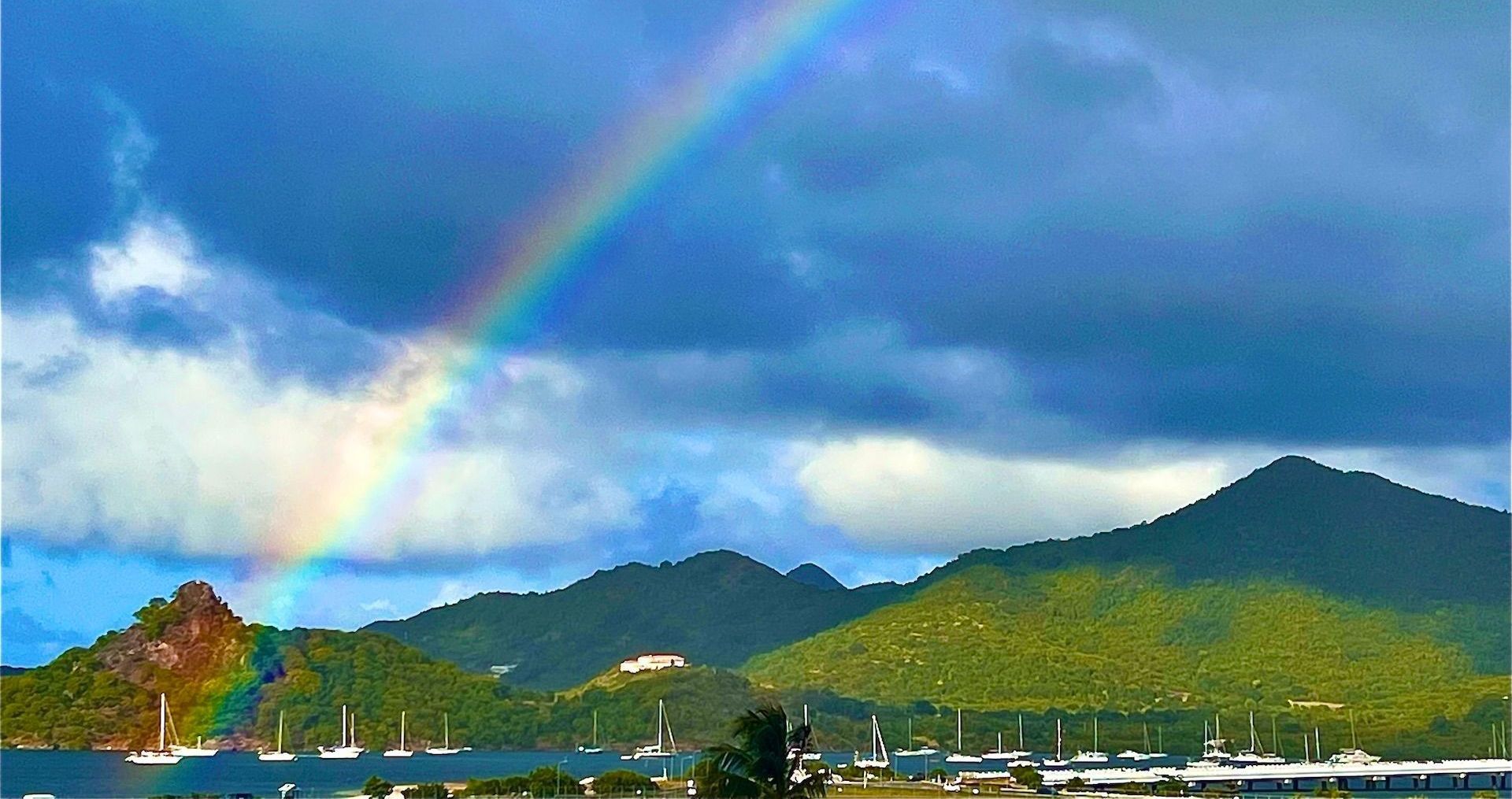 Rainbow arcs over green mountains and harbor with boats on a cloudy day.