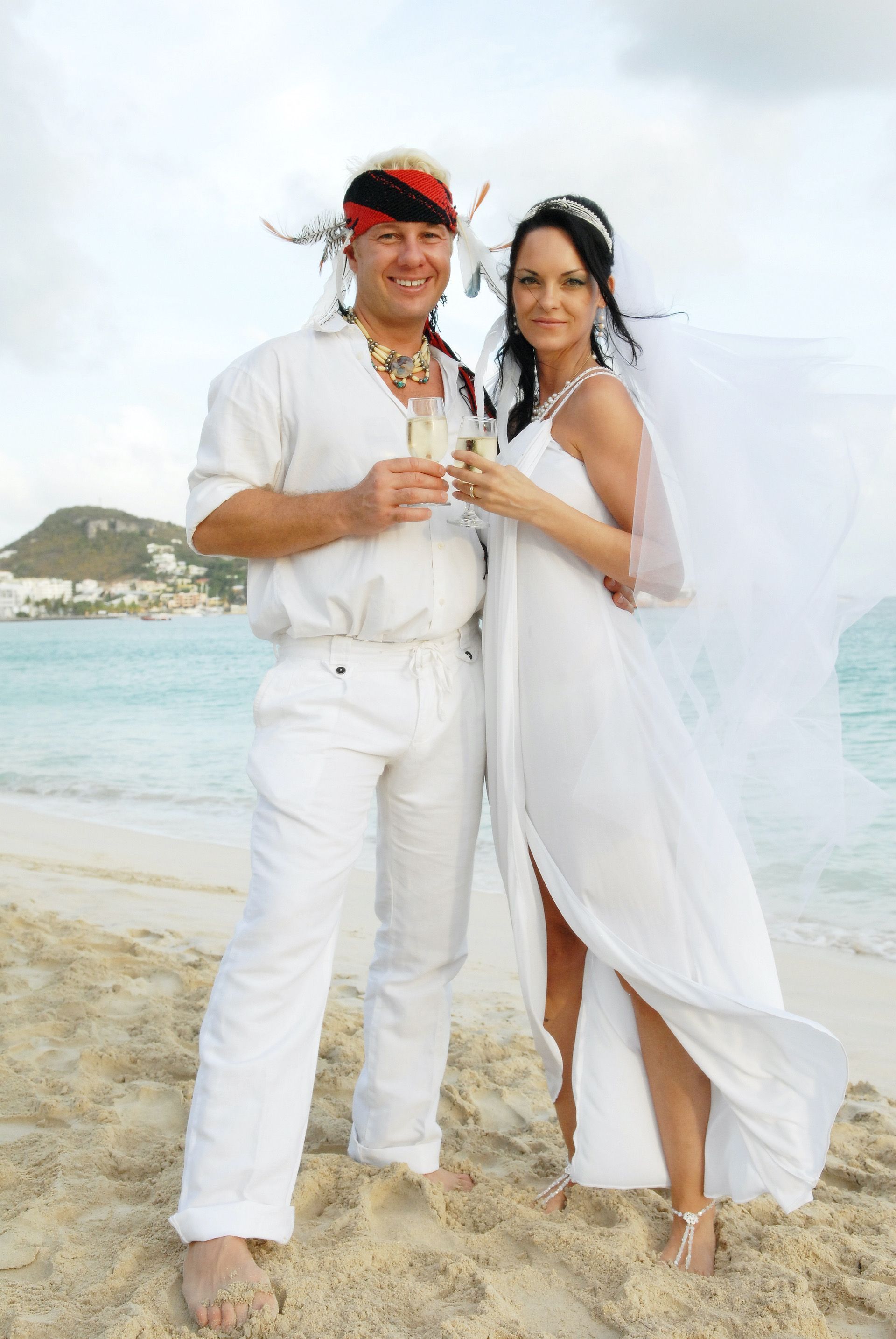 Couple in wedding attire holding champagne on a beach; woman in white dress with veil, man in white shirt and pants.