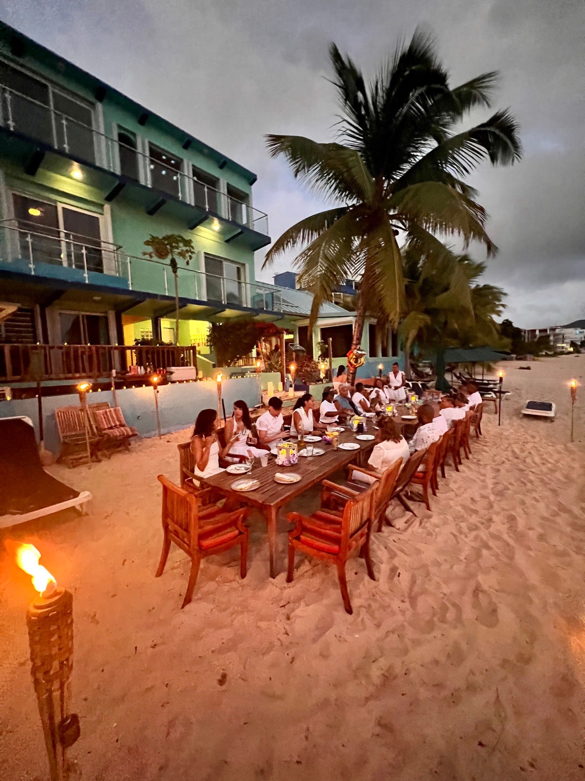 Beachside dinner party with long table, people eating, torches, and turquoise building at dusk.