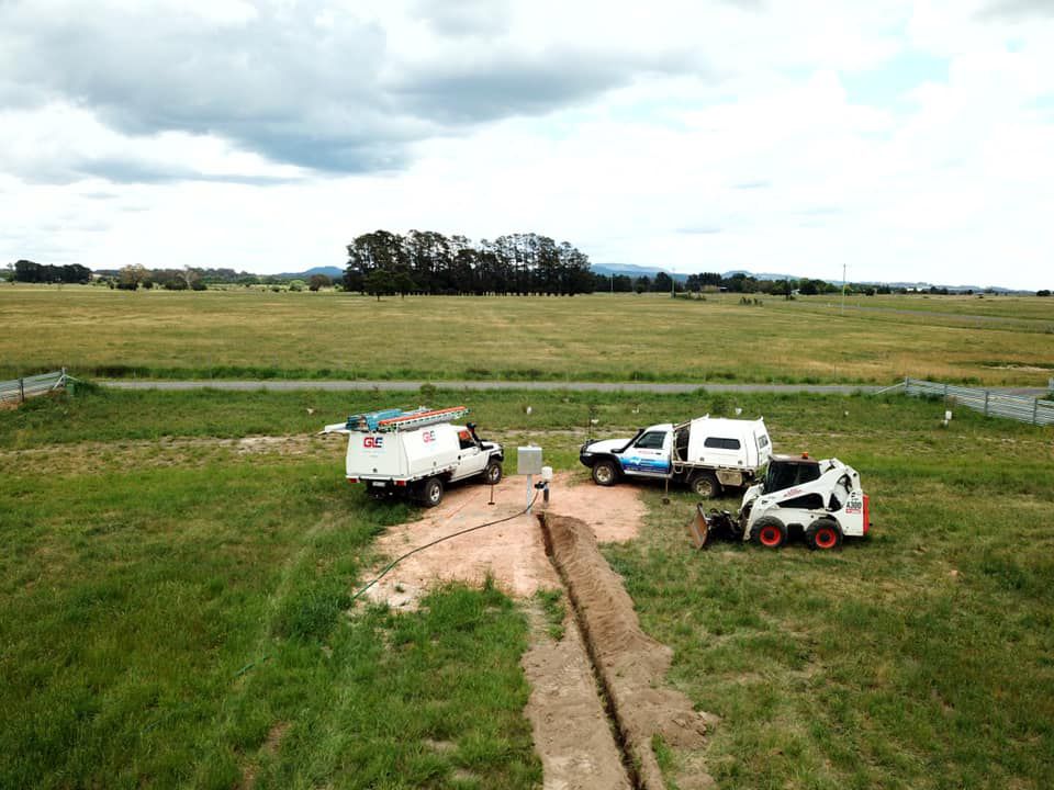 Workers and trucks in a grassy field beside a dirt track under a cloudy sky