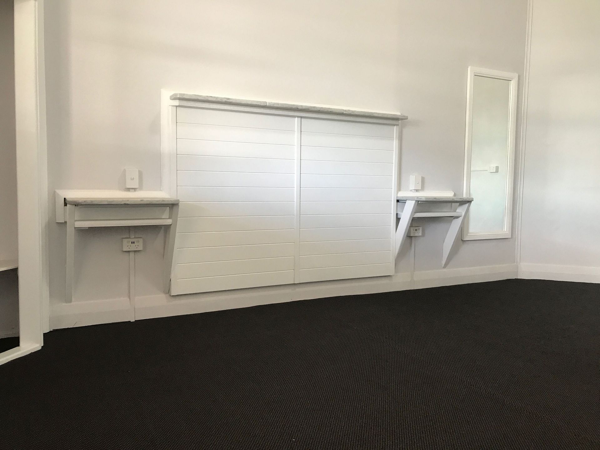 Empty room with white walls, shuttered window, small shelves, and dark carpet flooring.