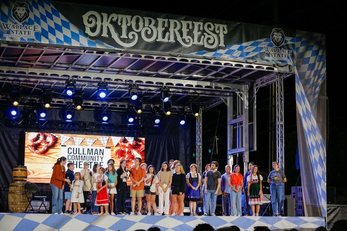 People on stage at Oktoberfest, a festival with a blue and white banner. A large screen displays food.