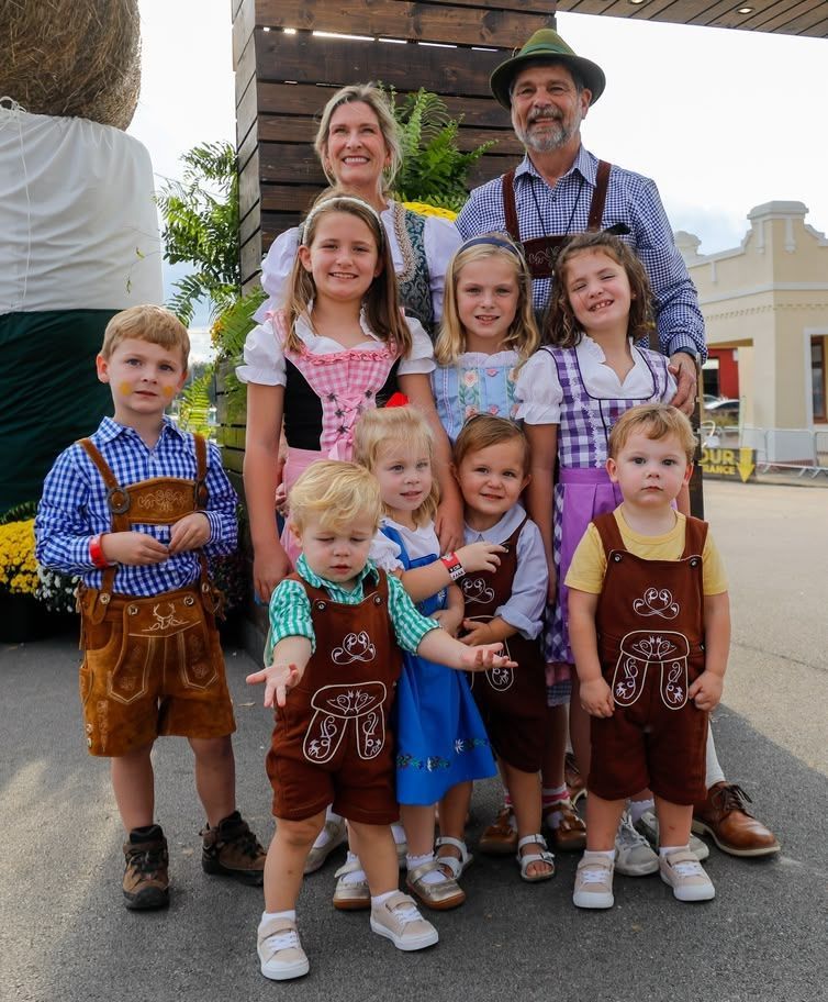 Large family posing outdoors in traditional Bavarian-style outfits, smiling in front of a building.