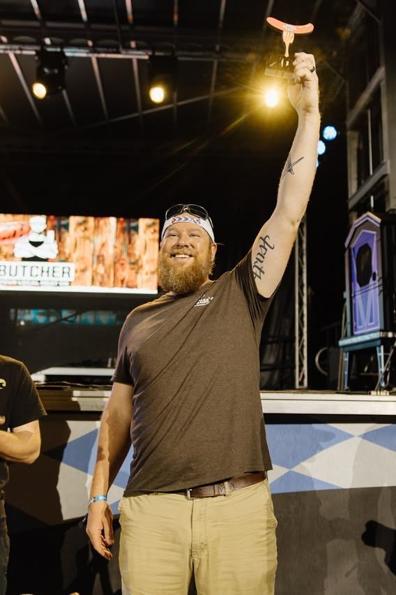 Man on a stage raises one arm under bright lights, wearing a dark shirt and tan pants.