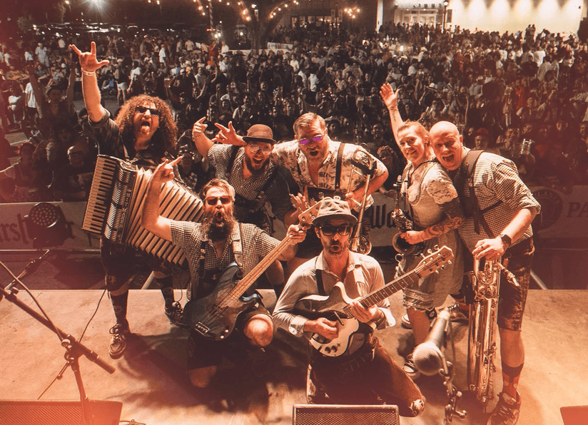 Band performing onstage, posing with instruments before a cheering crowd in warm orange light.