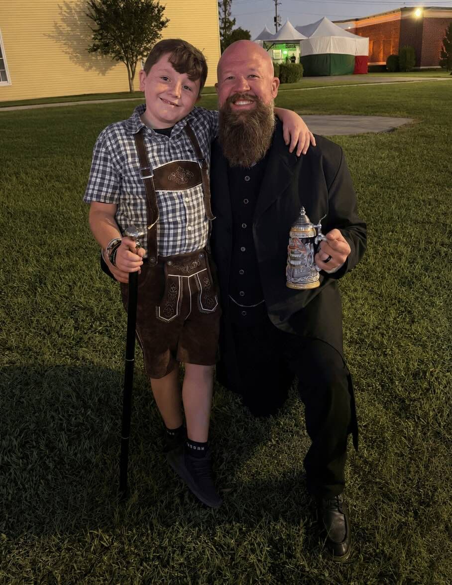Adult and child kneel together outdoors at night, smiling and holding drinks after an event.