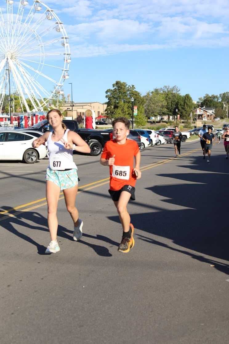 Two runners race past a Ferris wheel on a sunny street, with other people and cars in the background.