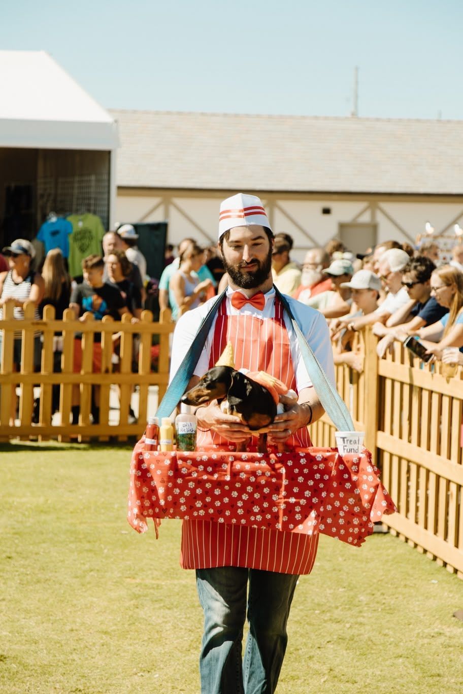 Man carrying a polka-dot basket at an outdoor event, with a crowd and wooden fence behind him