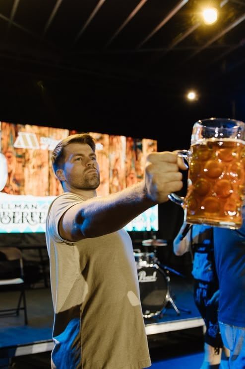 Man holding a large beer mug on a stage, with bright lights and a blurred crowd in the background