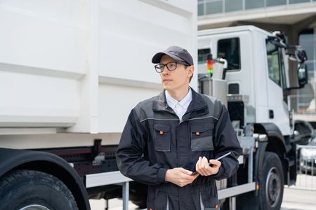 Worker standing beside a white truck, holding a device and wearing a dark uniform and cap.