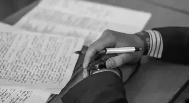 Hand holding a pen over open papers on a desk, with a cup nearby