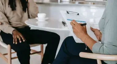 Two people seated at a table in a meeting, one holding a clipboard and pen near a coffee cup.