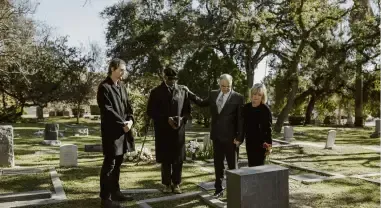 Four people stand among gravestones in a cemetery, appearing to hold a solemn outdoor memorial service.