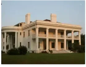 Large white neoclassical mansion with tall columns and a wraparound porch on a lawn