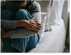 Person sitting curled up on a bed, hugging their knees in a soft gray sweater and blue jeans.