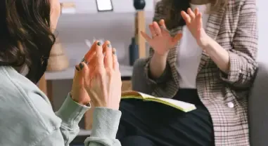 Two people talking in a meeting, one gesturing while the other listens with hands clasped.