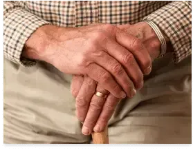 Hands clasped over a cane, with a gold ring and plaid shirt sleeves visible.