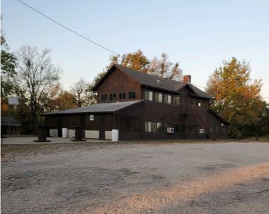 Dark wooden building with a sloped roof beside a gravel lot and autumn trees at dusk