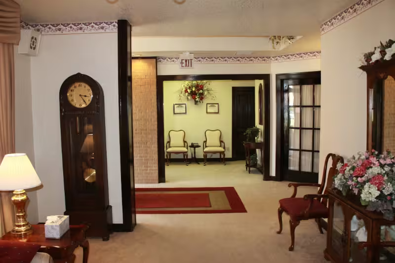 Elegant living room with grandfather clock, lamp, chairs, and floral arrangement leading into a hallway