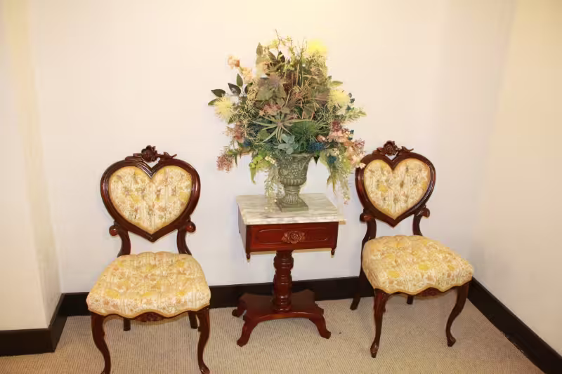 Two ornate upholstered chairs flank a small wooden table with a flower arrangement in a corner.