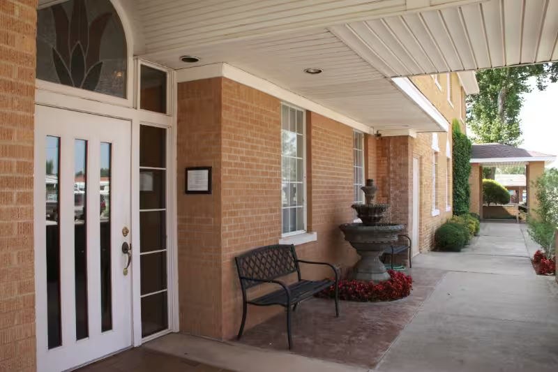 Brick building entrance with white door, bench, fountain, and concrete walkway under an overhang.