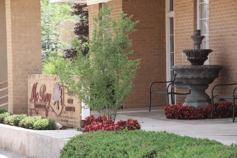Brick building entrance with fountain, red flowers, and a stone sign by the walkway