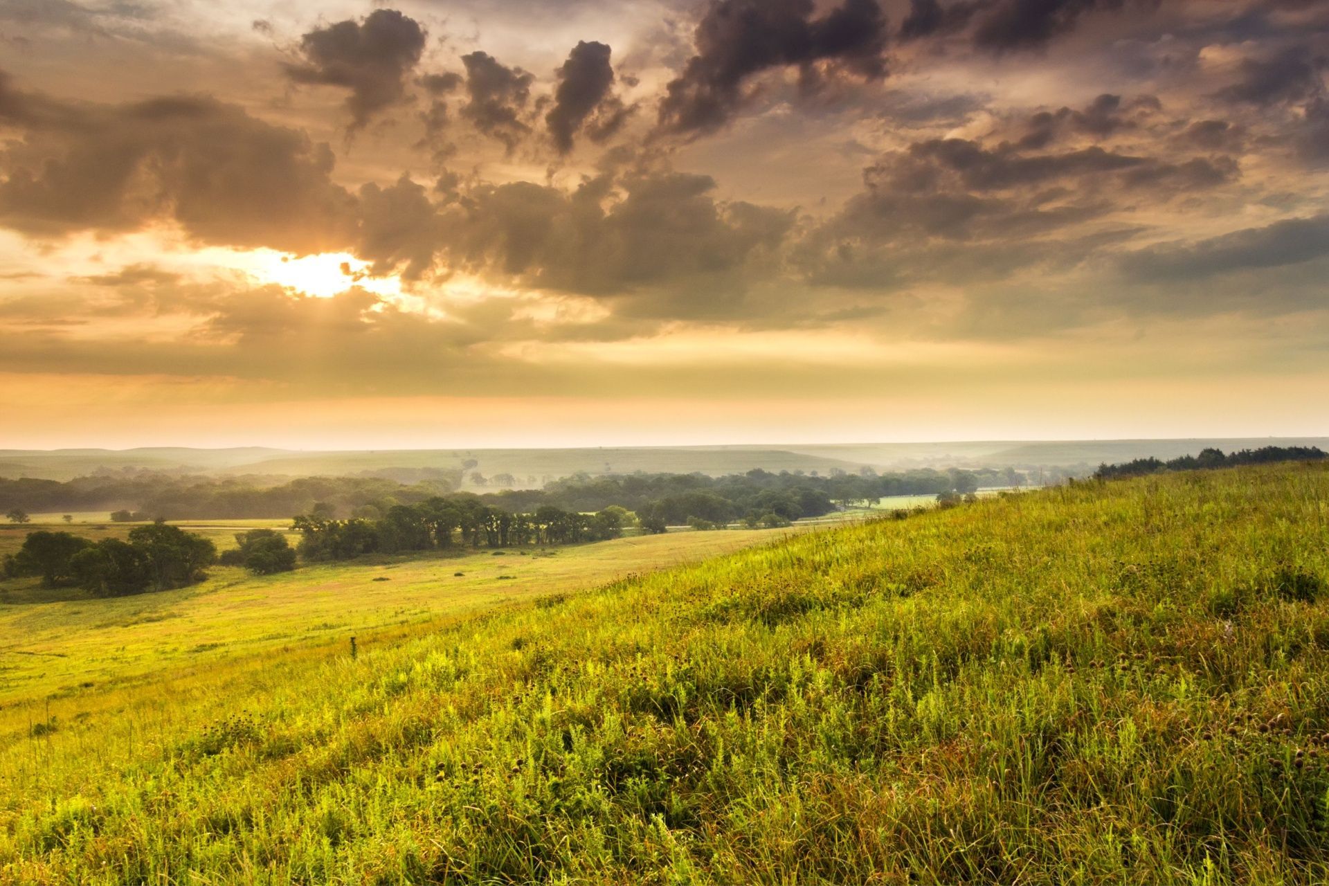 Sunlit grassy hillside overlooking a distant valley under dramatic clouds at sunset
